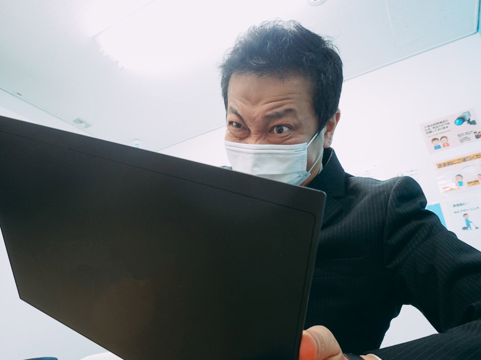 マスク姿でノートパソコンを持つ男性エンジニアが、オフィスで戸惑った表情を見せている［モデル：のせさん］