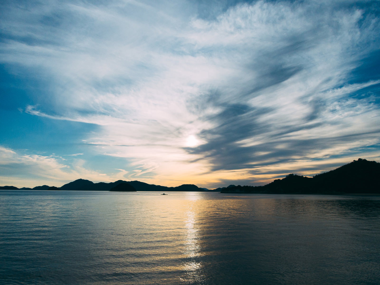 夕焼け空の下で海面に夕日が反射し、遠くに山のシルエットが見える風景