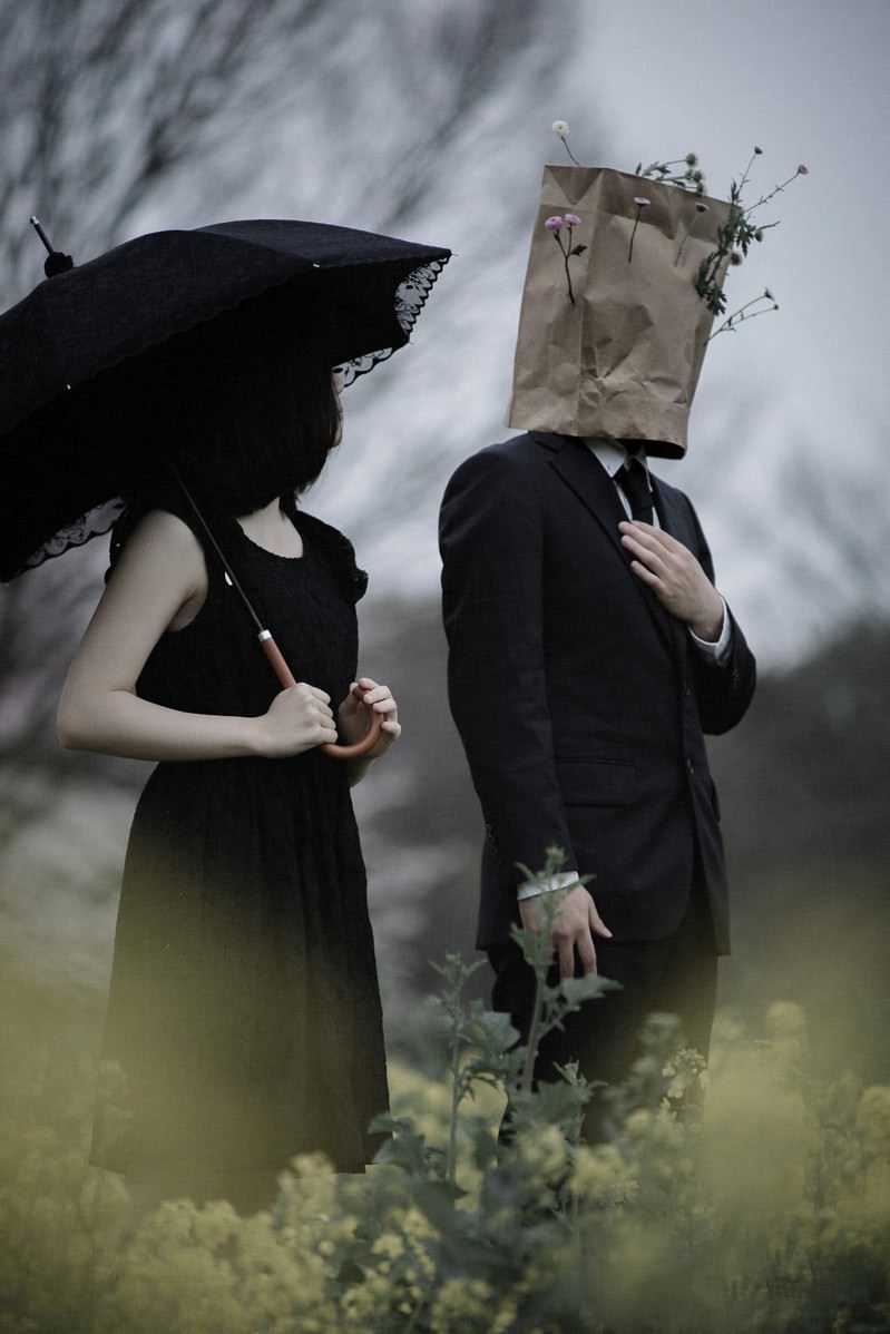 菜の花畑で向かい合う紙袋を被った男性と黒い服の女性