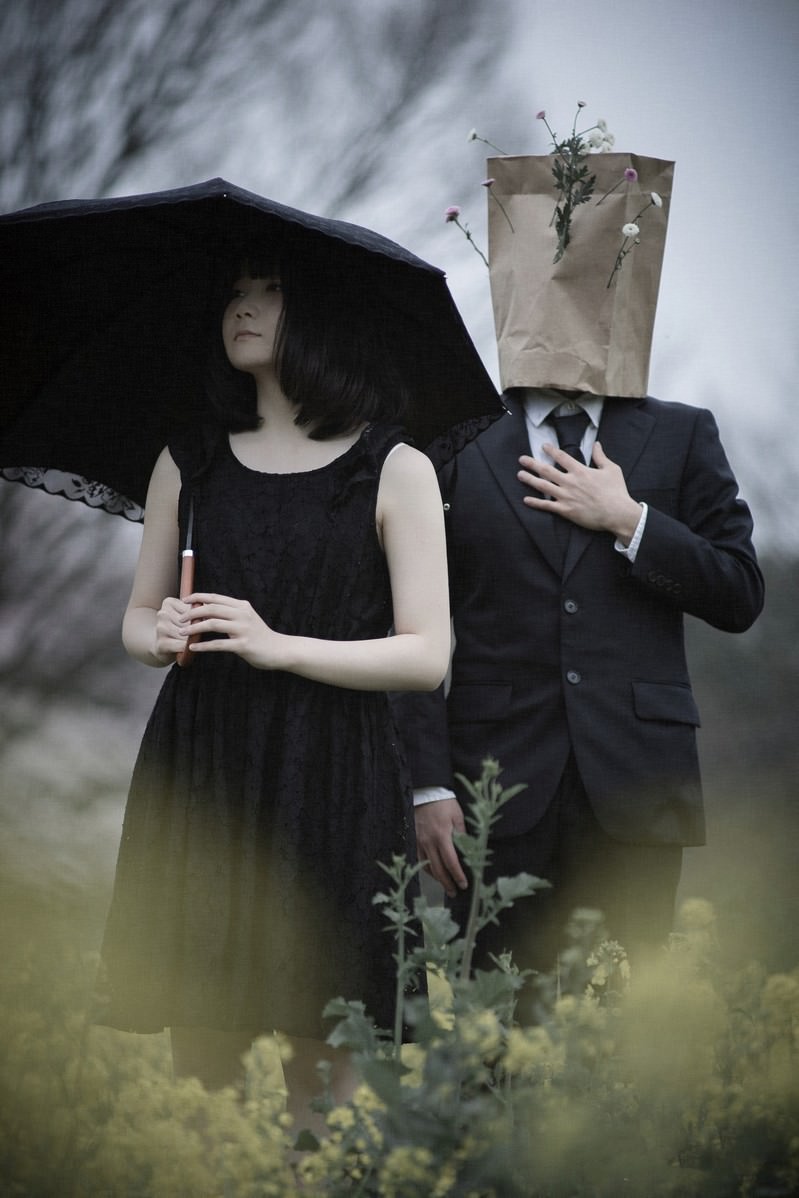 紙袋を被った男性と黒いドレスの女性が菜の花畑に立つ