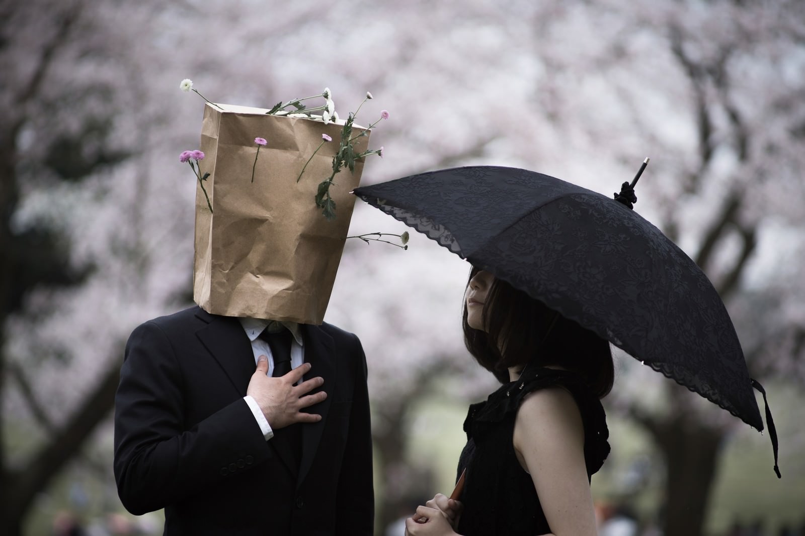 桜の背景で紙袋を被った人と黒い傘を持つ女性が並んで立っている