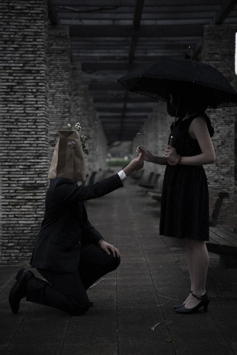 薄暗い石造りの建物内で黒い衣装の男性が跪いて女性に花束を渡している