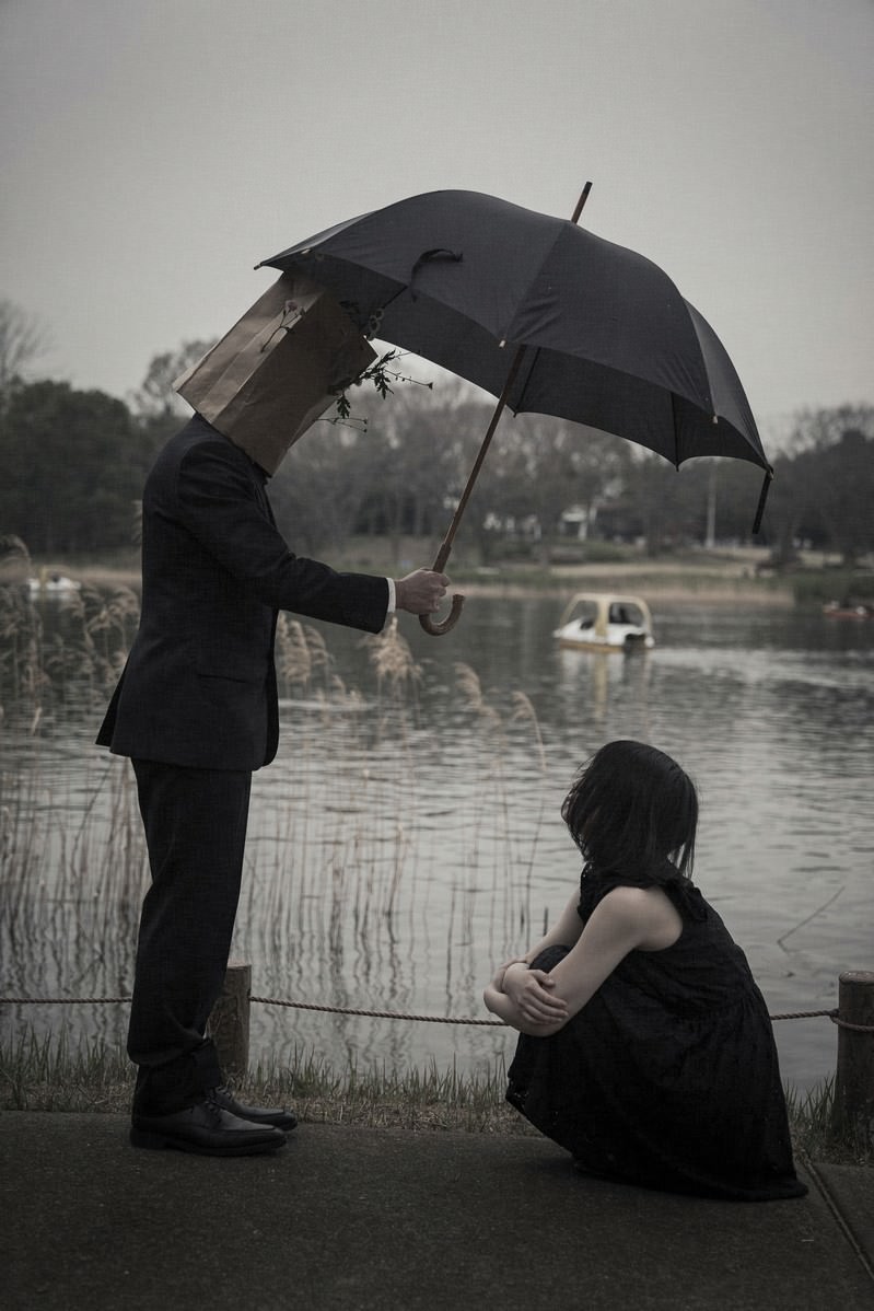 雨の中で座り込む女性に傘を差し出す男性のモノクロ写真