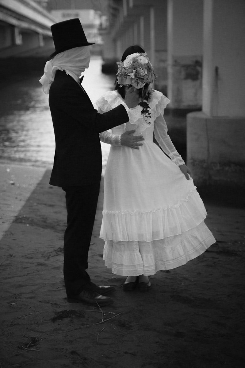 覆面をした男性がブーケを掲げ、白いドレス姿の女性と並んで立つモノクロ写真
