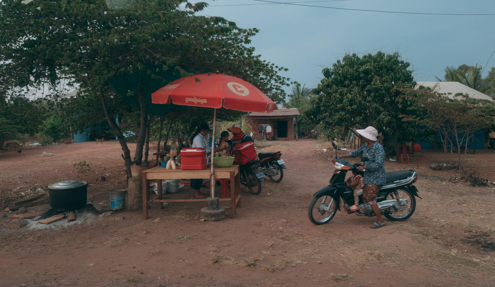 A red parasol stall set up on a dirt road in Cambodia, with local residents riding scooters passing by.
