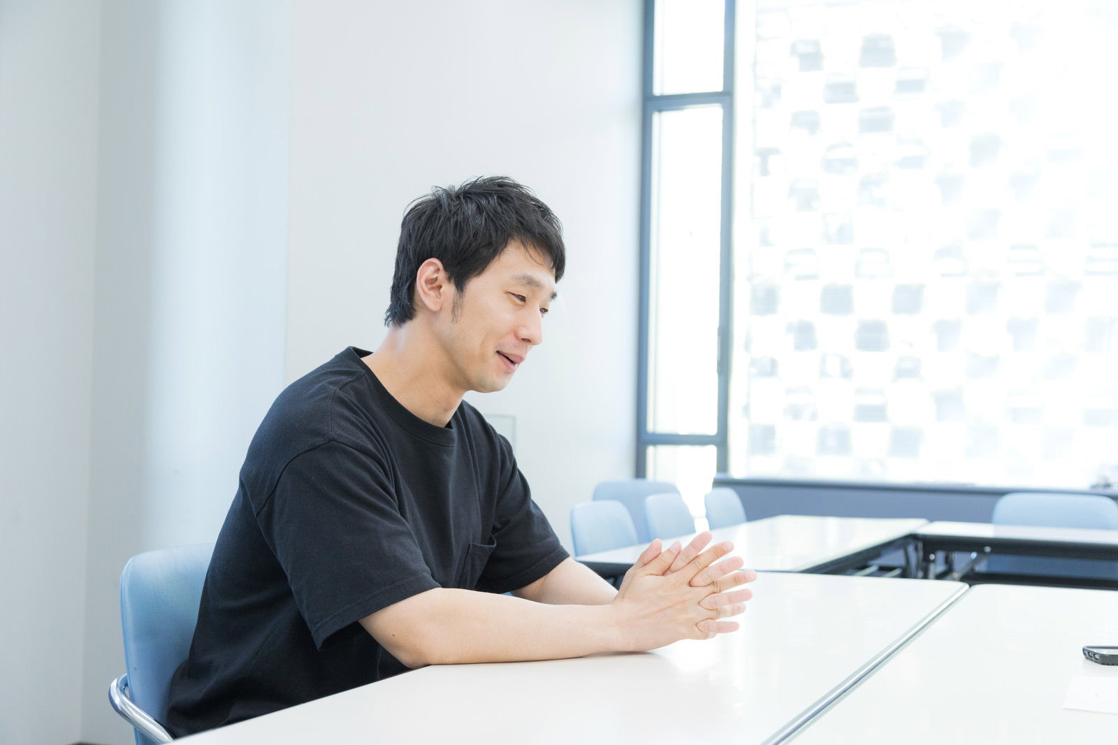 明るいオフィスのデスクで話をしている黒いシャツの男性［モデル：大川竜弥］