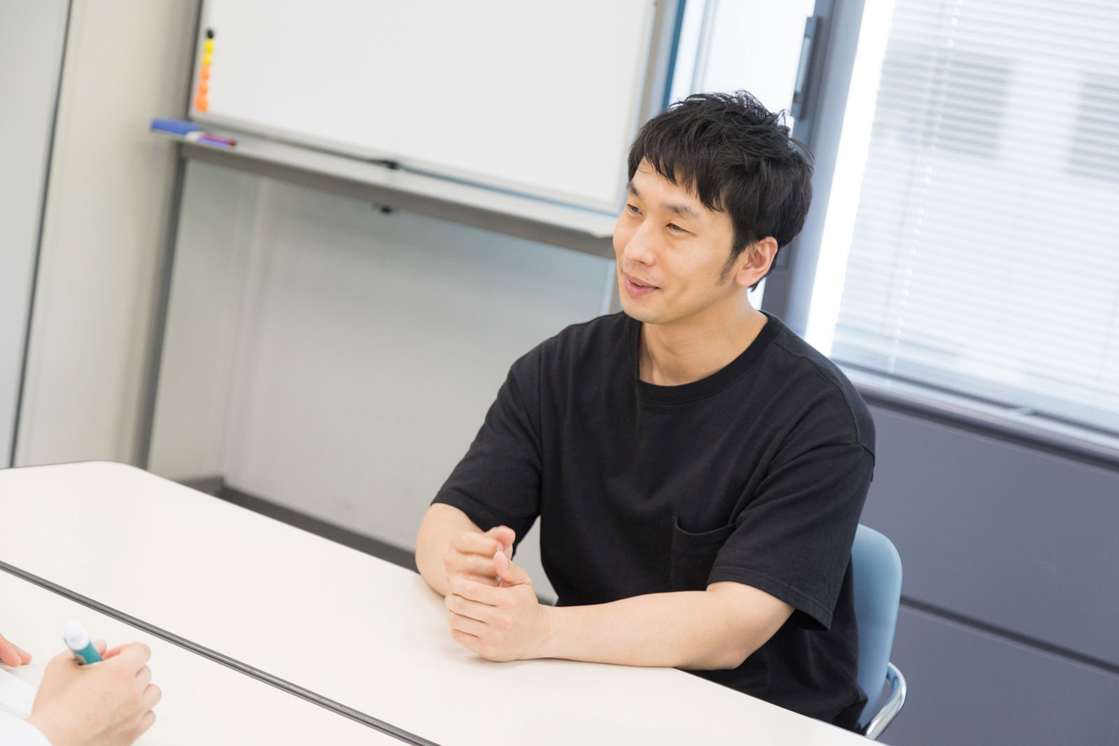 黒いトップスを着た男性が椅子に座り手を組んで話している［モデル：大川竜弥］
