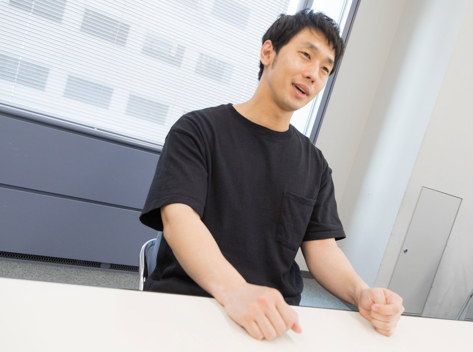 黒いTシャツを着た男性が机に手を置いてインタビューに答えている様子［モデル：大川竜弥］