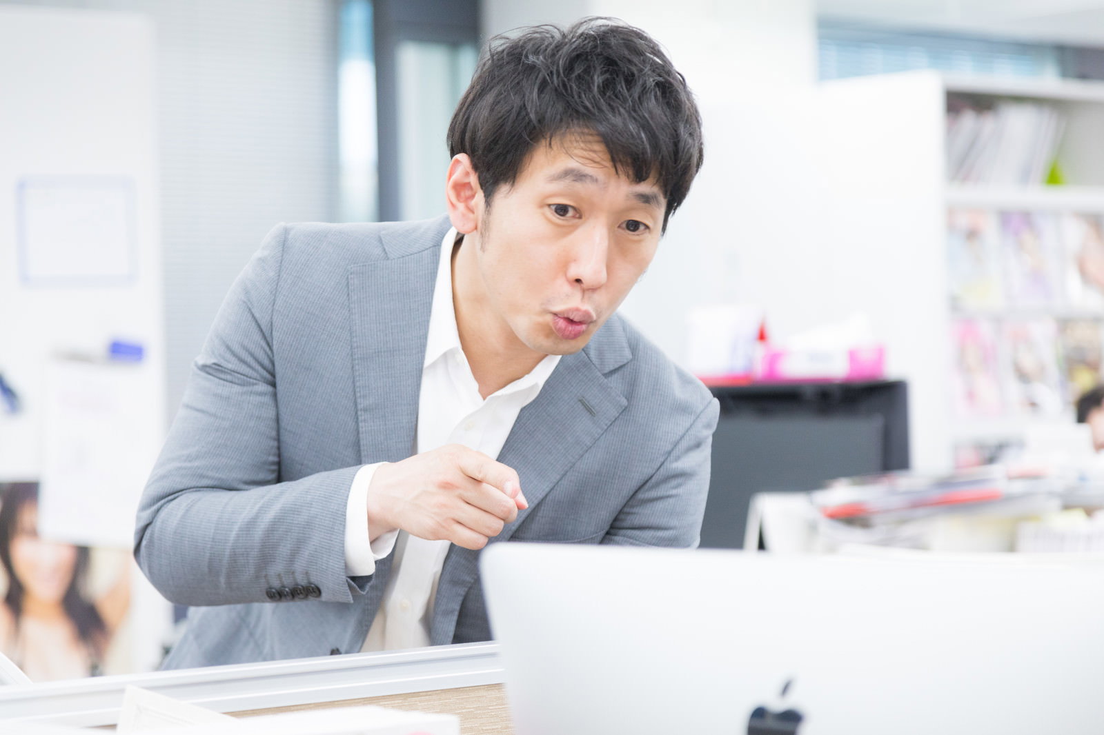 グレーのスーツを着た男性会社員がオフィスのデスクで人差し指を立てて指摘している［モデル：大川竜弥］