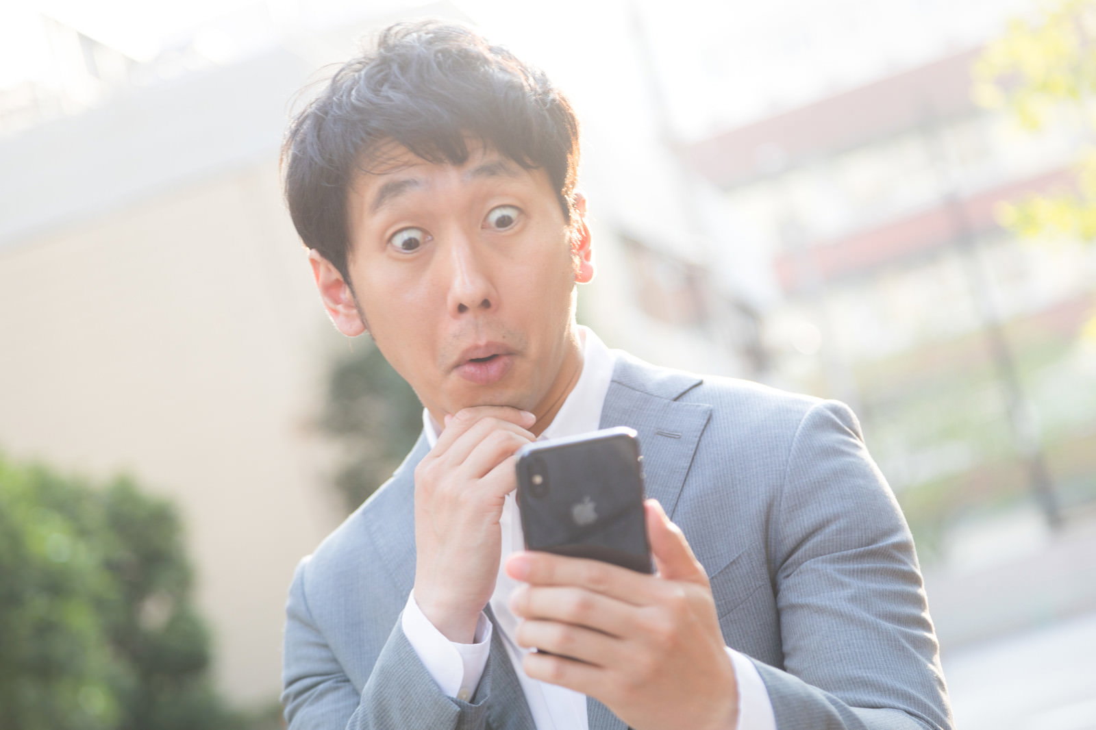 スマートフォンを手に持ち驚いた表情を浮かべるグレーシャツの男性［モデル：大川竜弥］