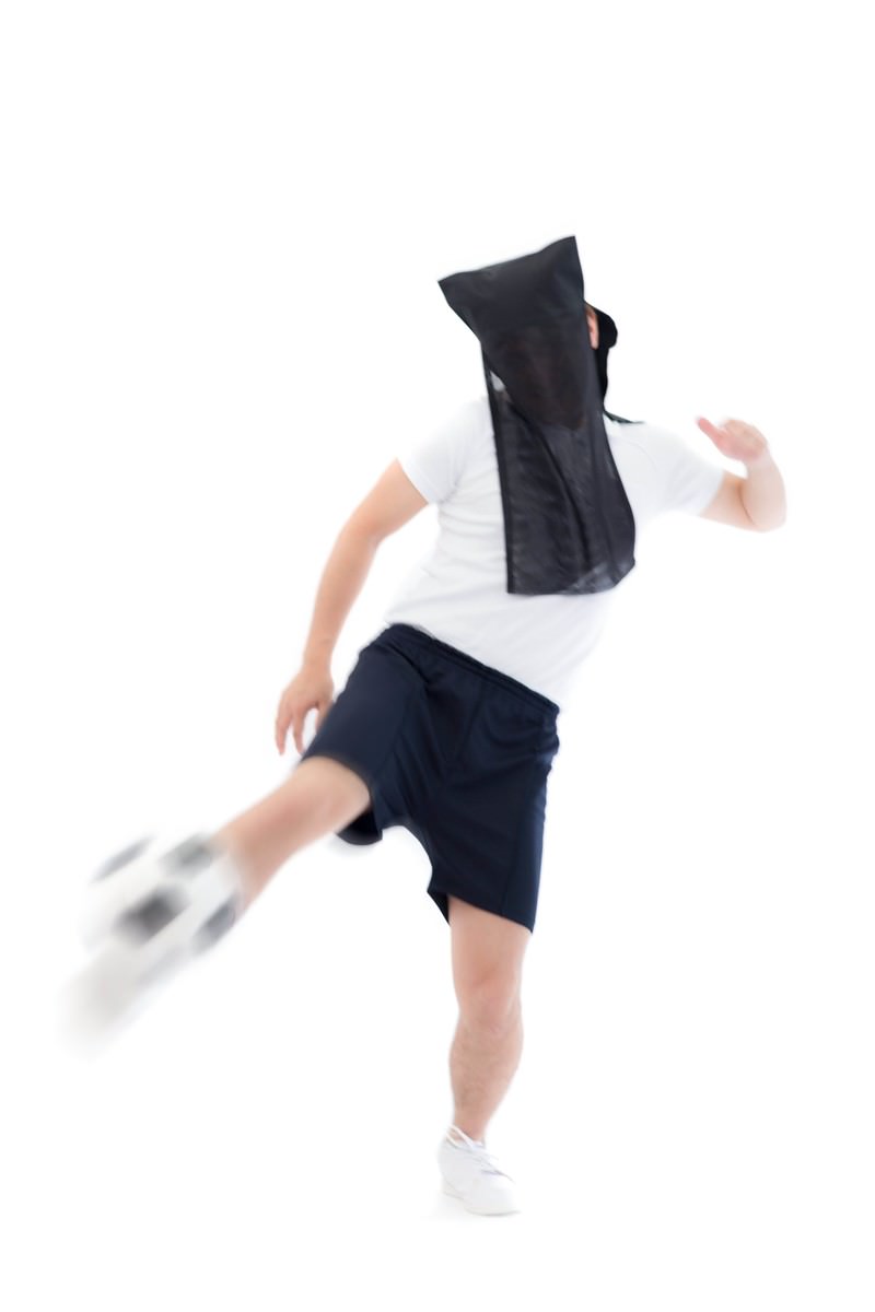 黒い布で顔を覆った人物が右足を高く上げてサッカーボールをドライブキックしている動きのある場面［モデル：鈴木秀］