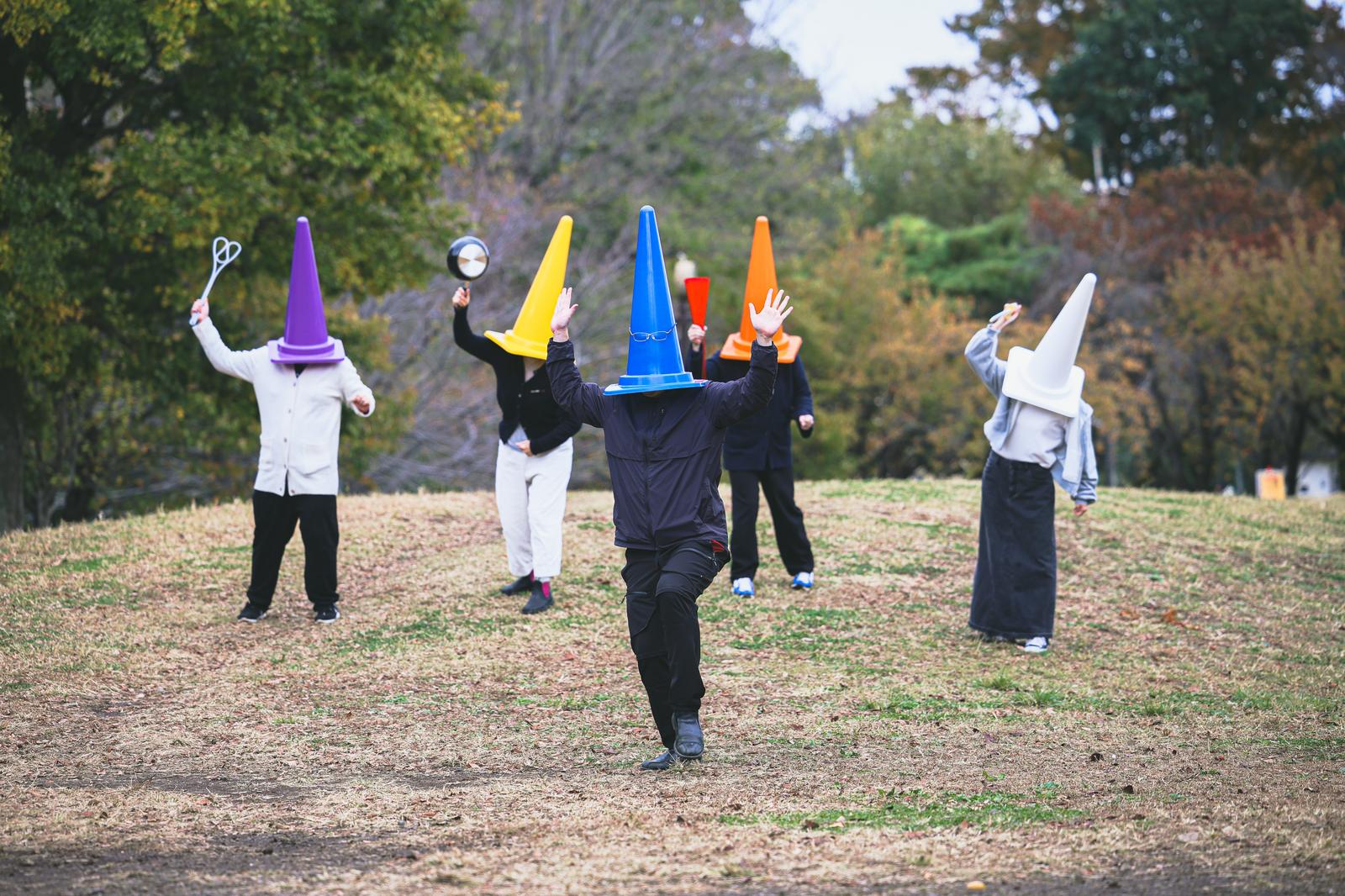 公園で青い三角コーンをかぶった人が、複数の色付きコーンをかぶった女性に追われている
