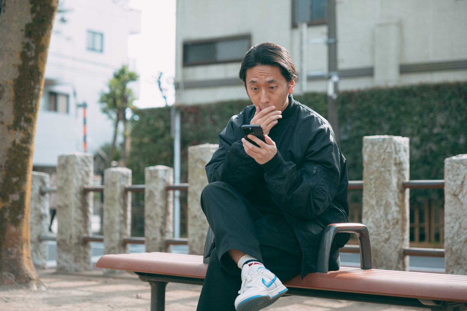 スマートフォンを見て驚く黒いジャケット姿の男性がベンチに座っている［モデル：大川竜弥］