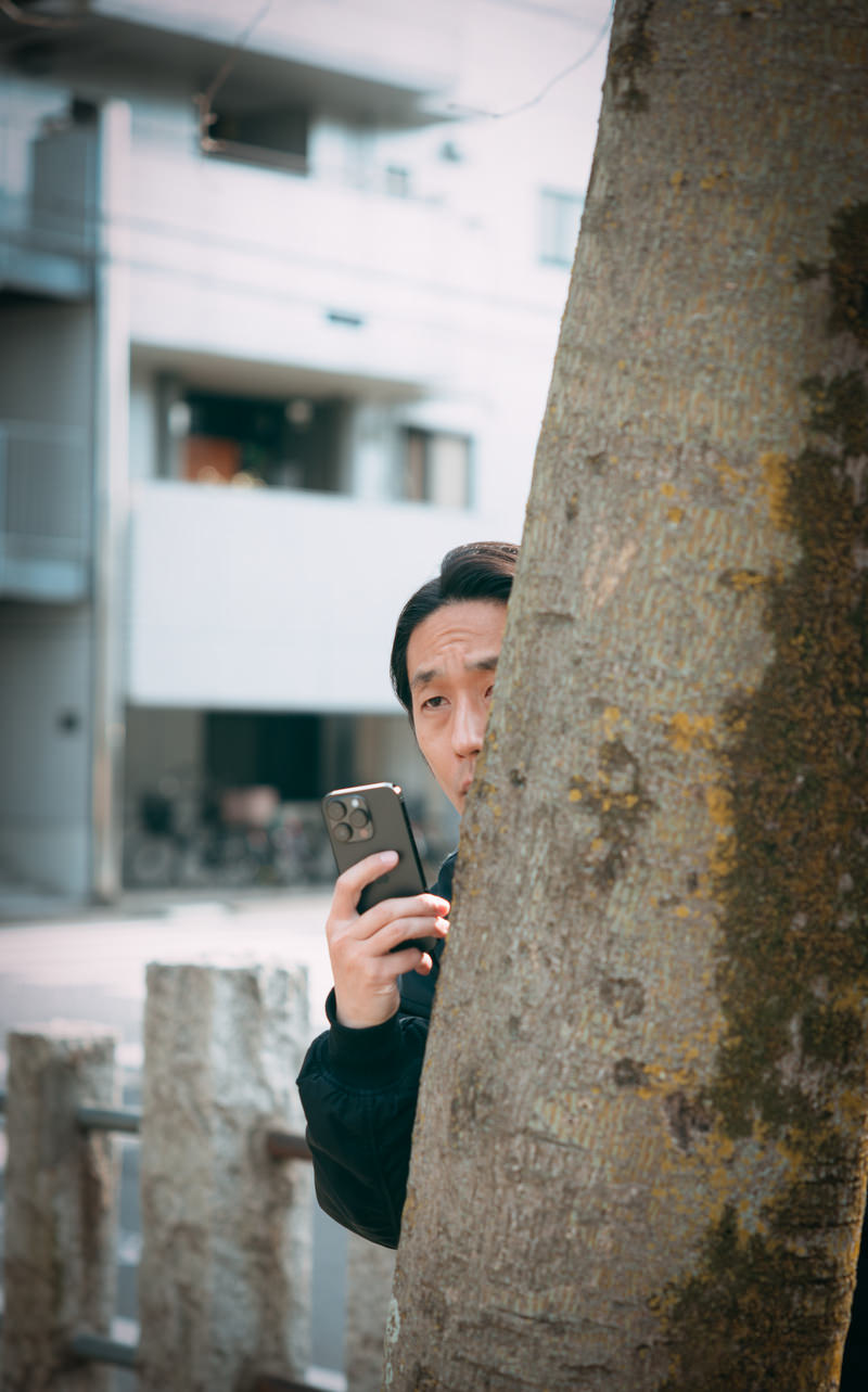 木の幹に隠れてスマートフォンで何かを撮影する男性［モデル：大川竜弥］
