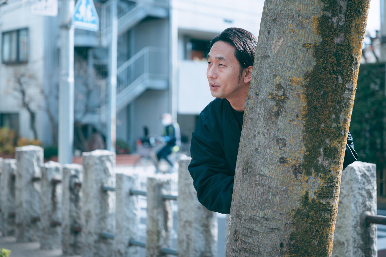 樹の幹の奥から身を隠して覗き見する黒い服の男性［モデル：大川竜弥］