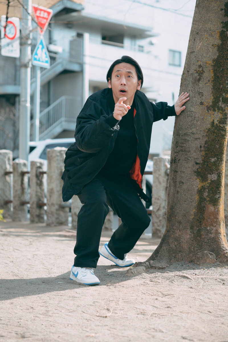 公園の樹木の前で驚いた顔をしている黒いジャケットの男性［モデル：大川竜弥］