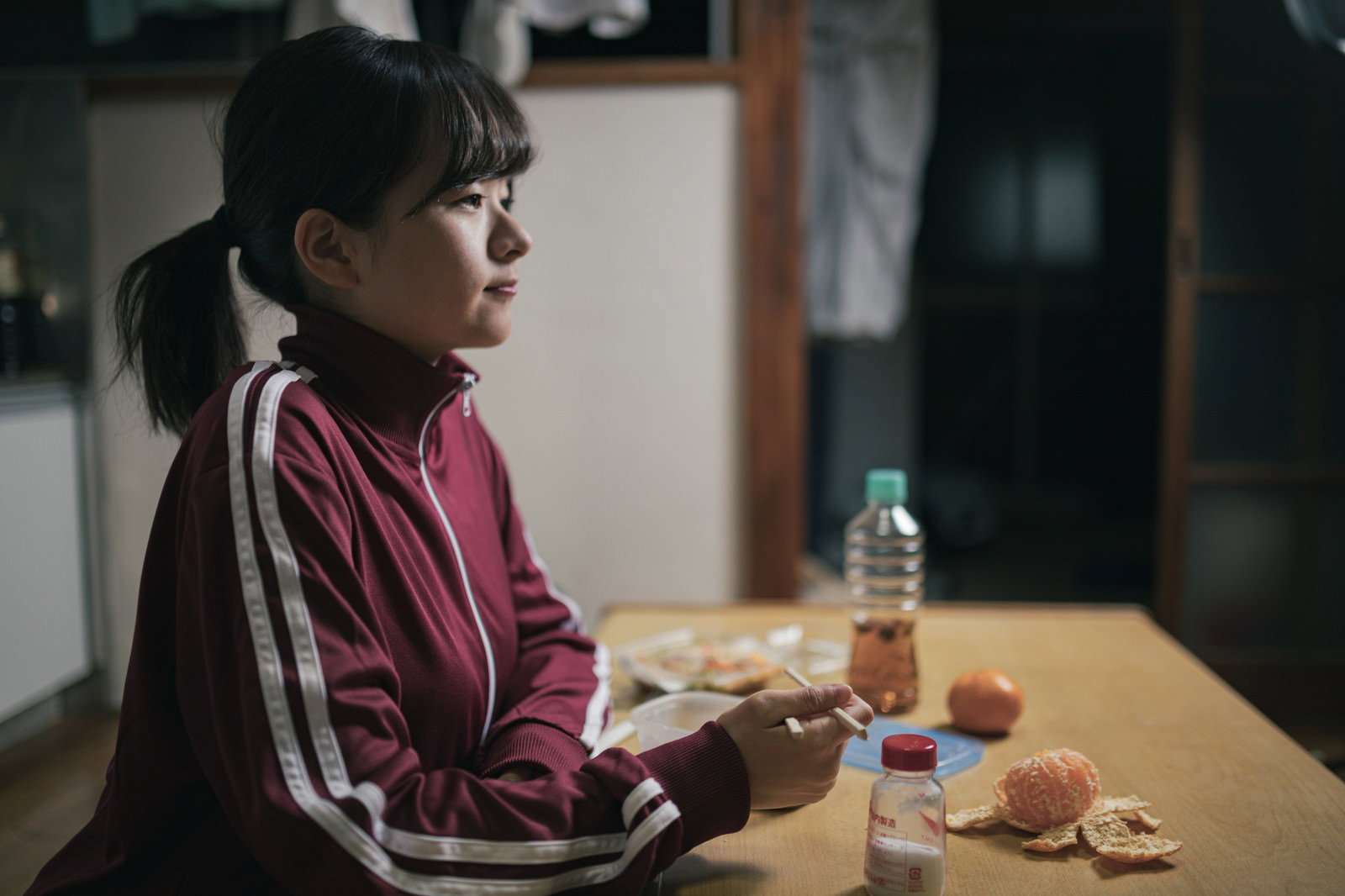 ジャージを着た若い女性が薄暗い室内の食卓に座る［モデル：千歳］