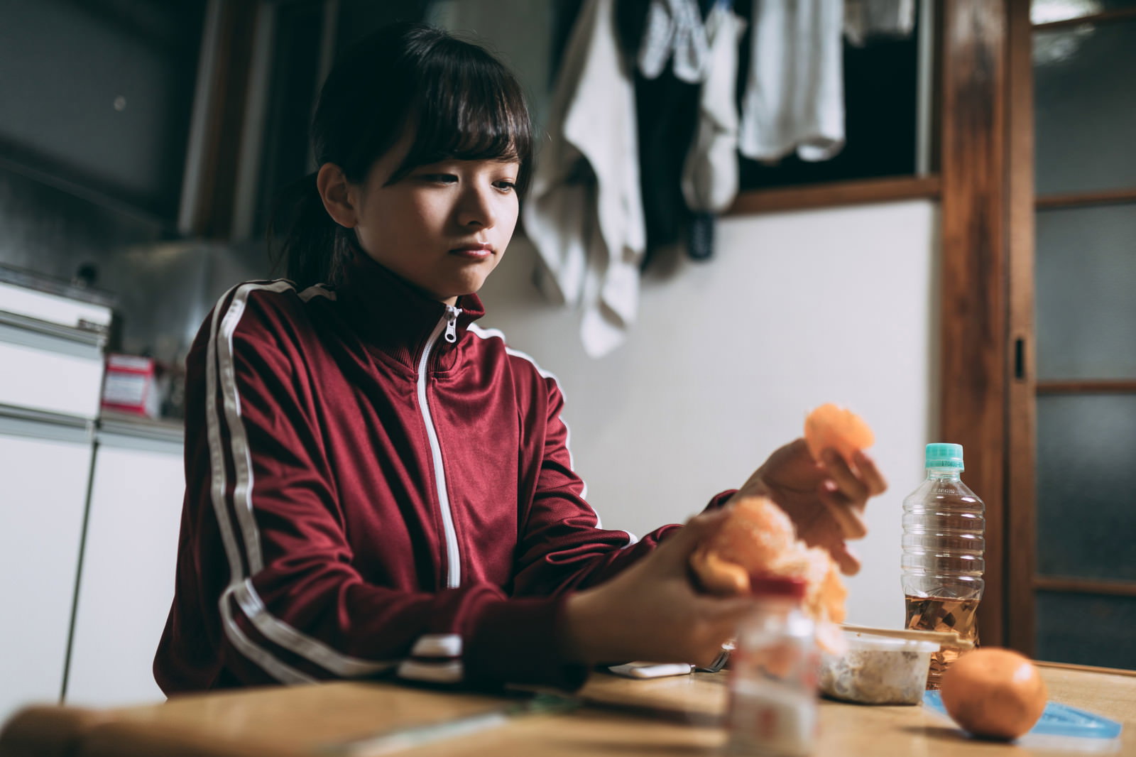 赤いジャージを着た女性がワンルームでみかんを持っている［モデル：千歳］