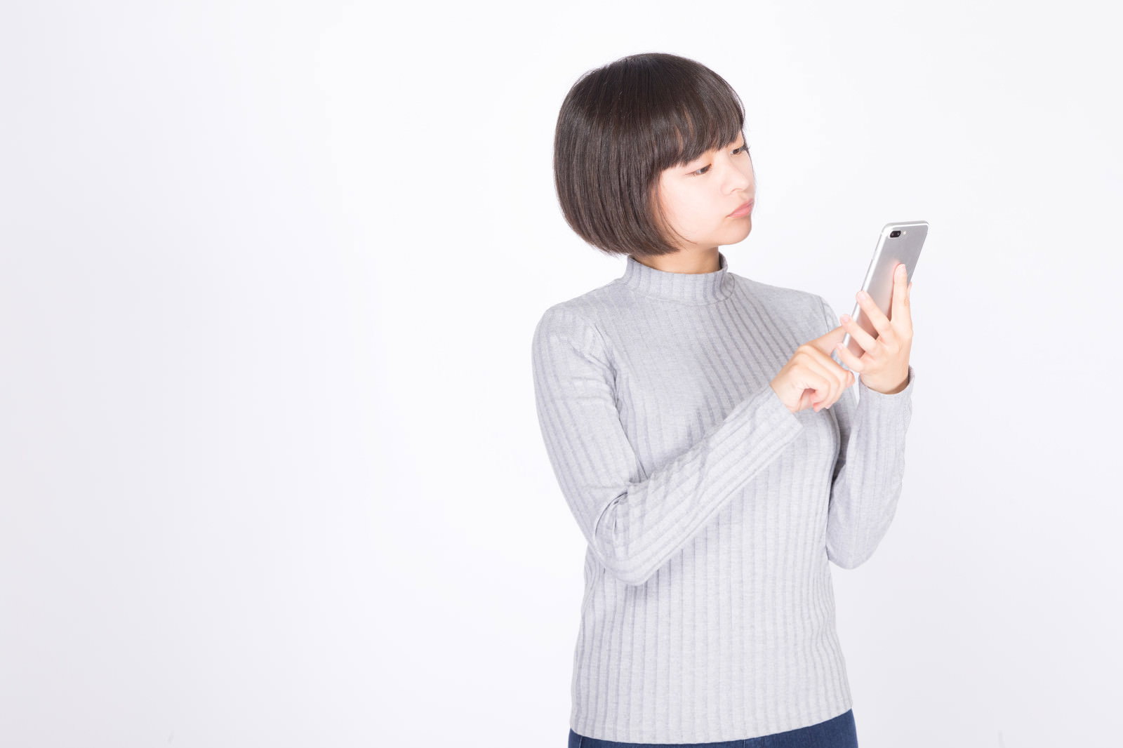 白い背景でグレーのニットを着た女性がスマートフォンを操作している［モデル：千歳］