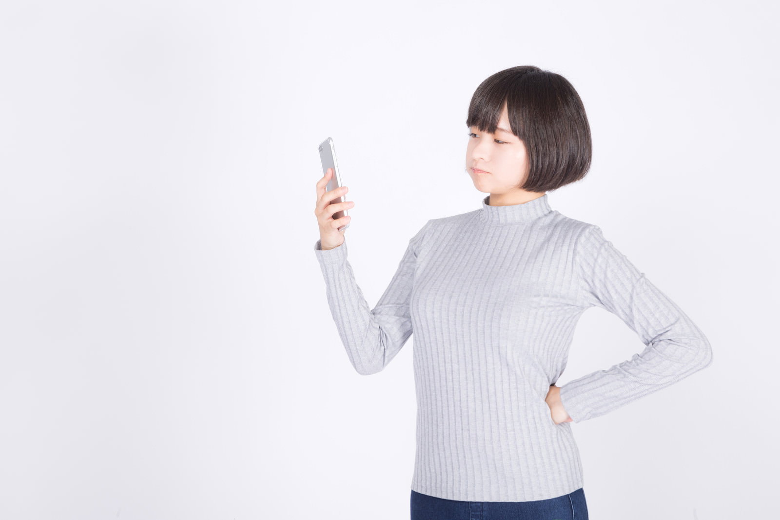 白い背景の前でスマートフォンを手に持ち呆れた表情を浮かべる女性［モデル：千歳］