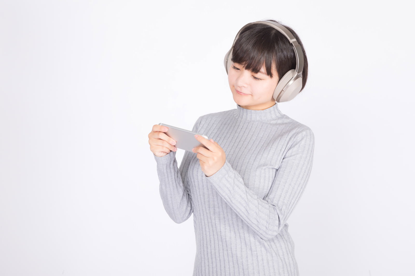 グレーのニットを着た女性がワイヤレスヘッドホンを装着してスマートフォンを操作している［モデル：千歳］