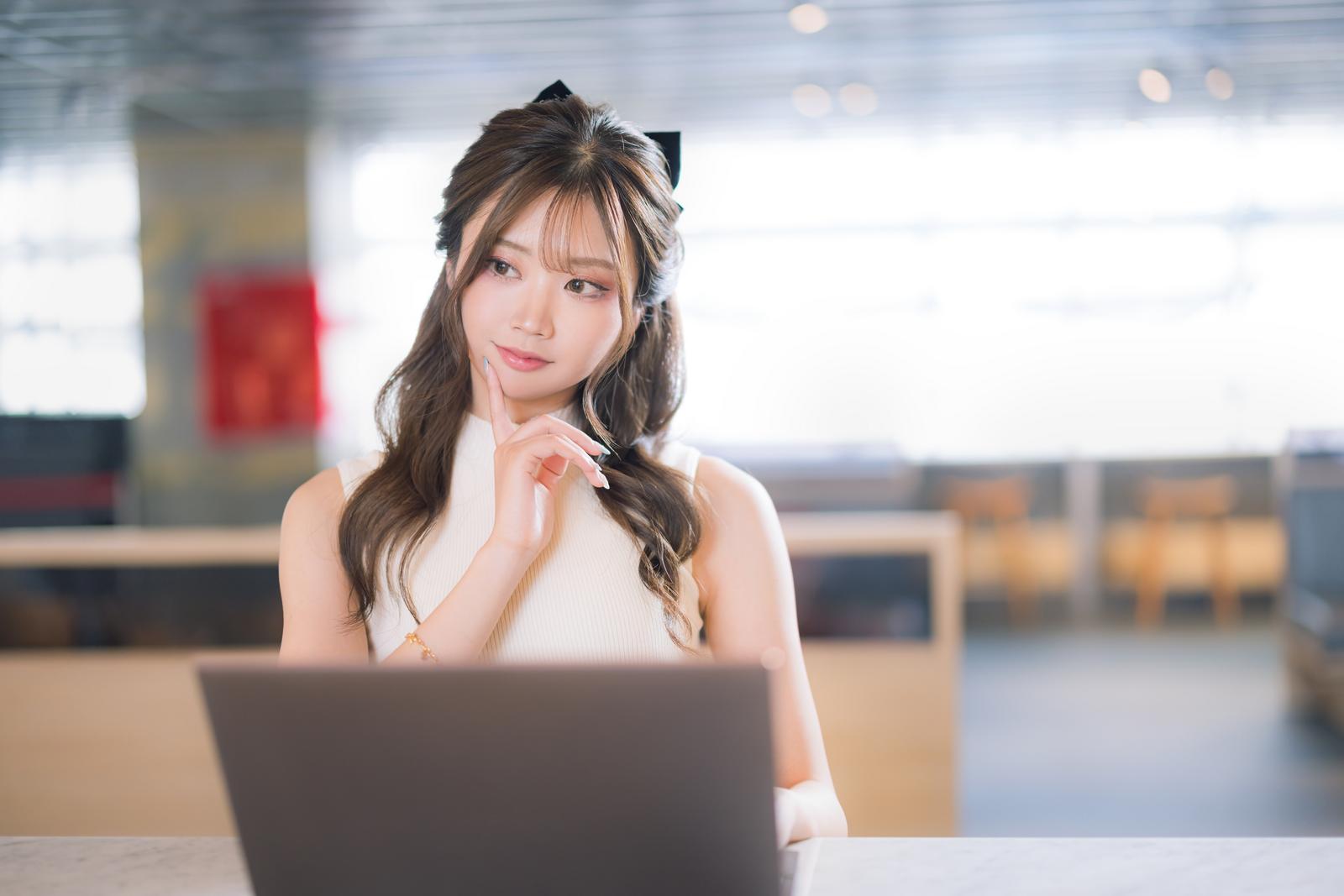 ノートパソコンの前で考えながら悩む女性［モデル：れいか氏］