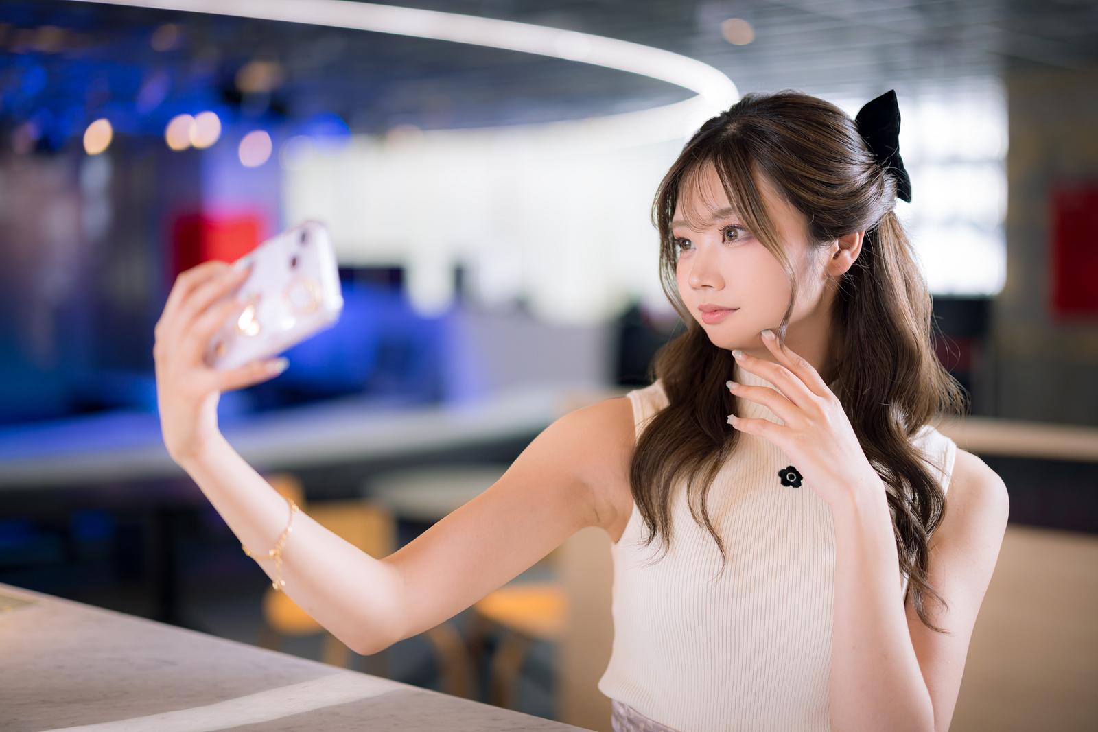 ネオン照明のあるカフェで自撮りをする黒いリボンヘアの女性［モデル：れいか氏］