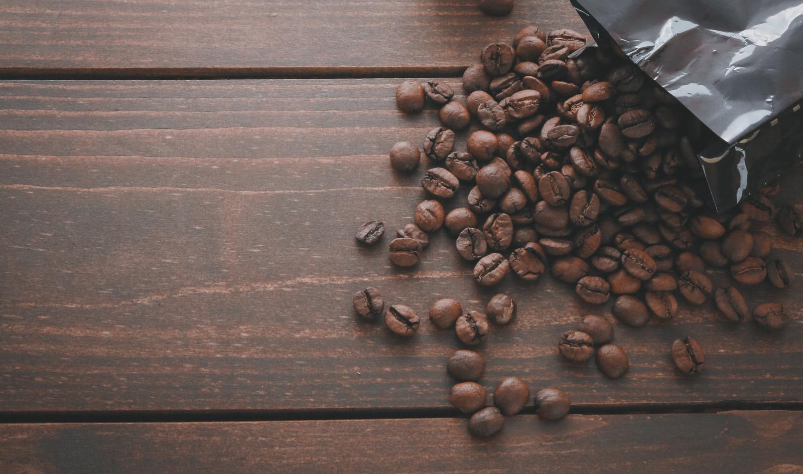 Roasted coffee beans spilling out of a black package and scattered across a wooden table