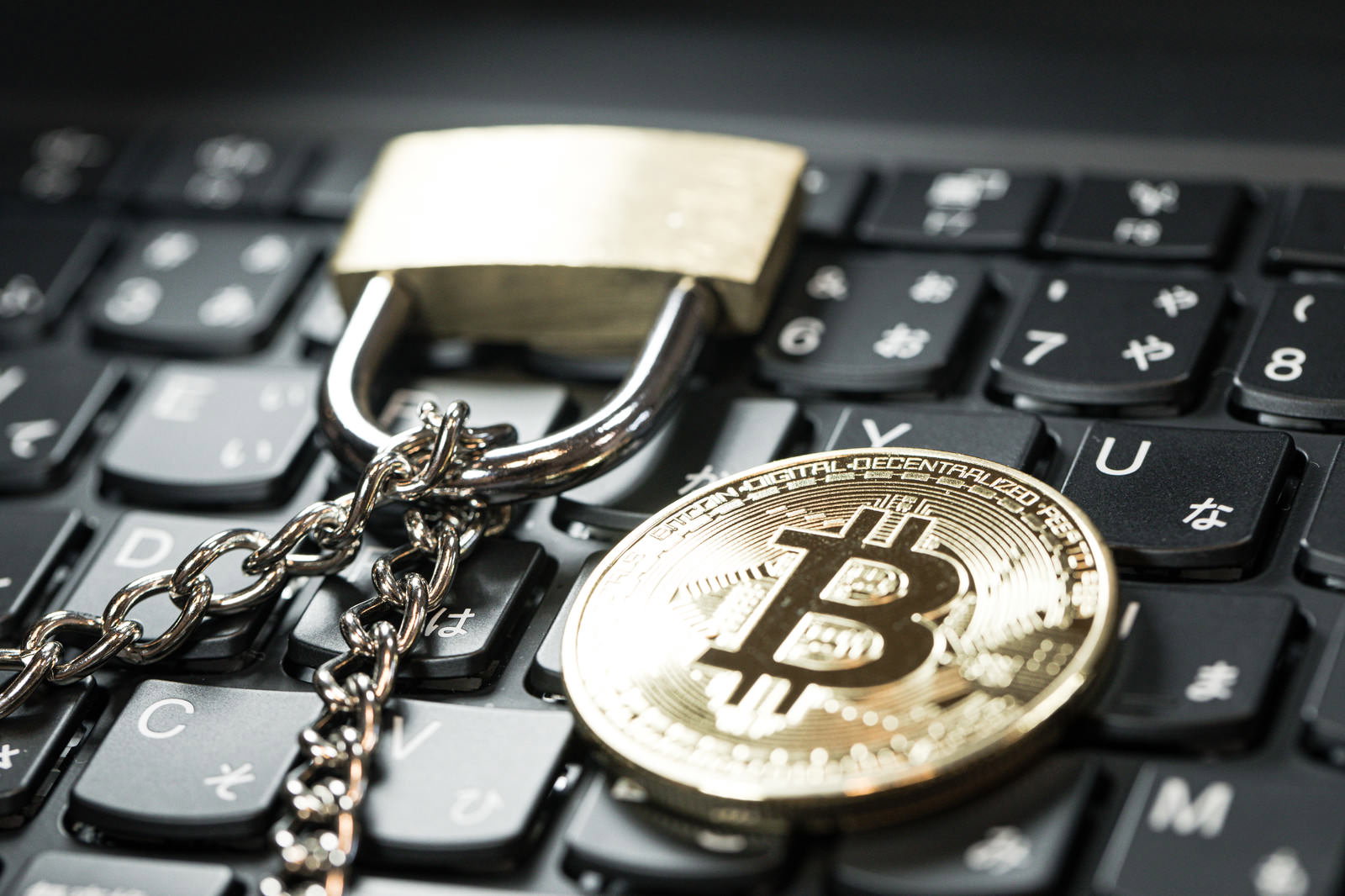 黒いキーボードの上に金色のビットコインコインと南京錠が鎖で繋がれている