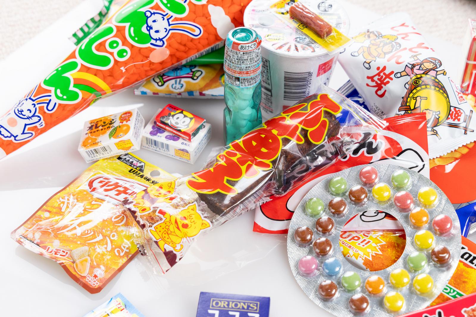 オレンジと緑のキャラクター入りパッケージの駄菓子やラムネ、グミが白い背景に広げられている