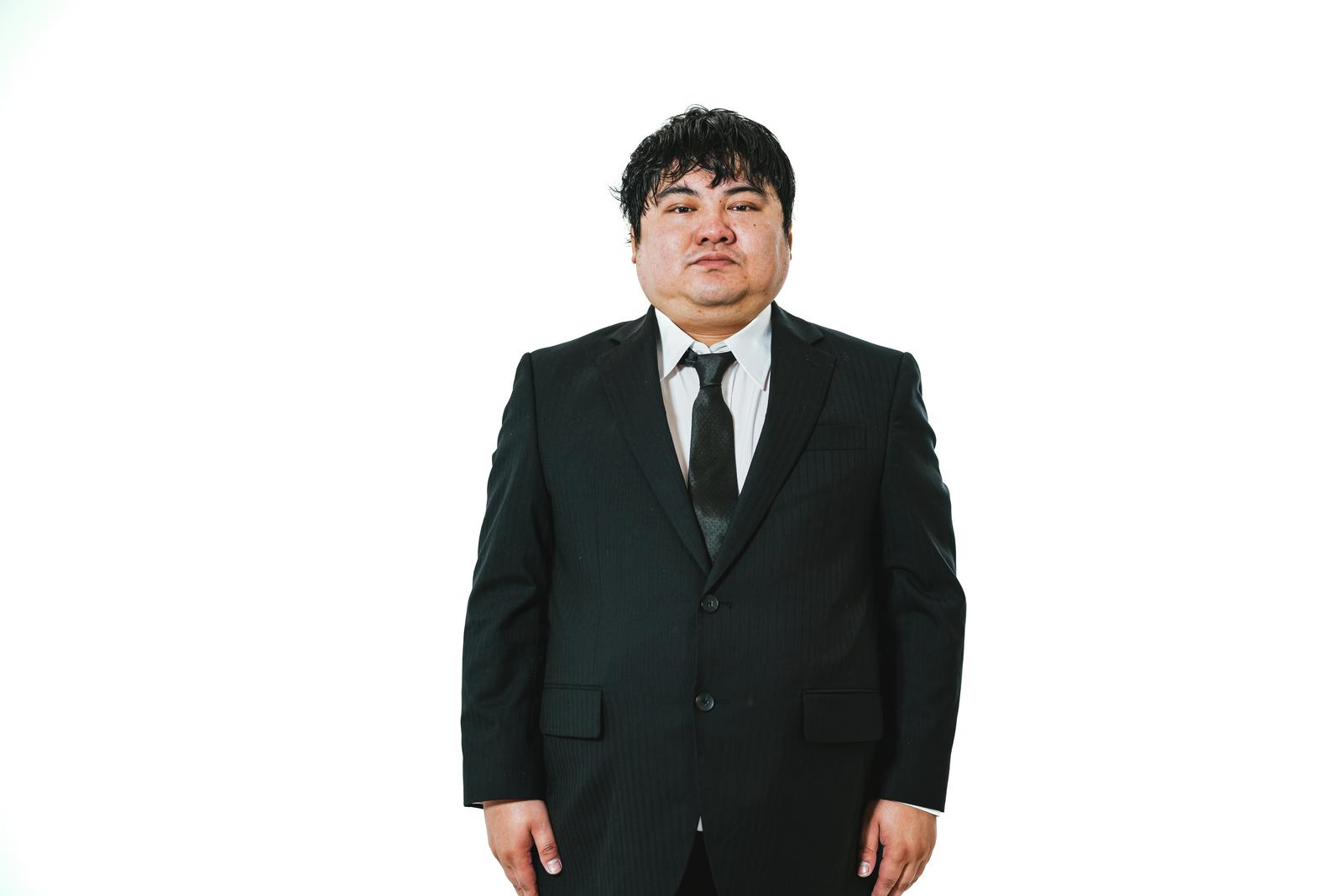 Stern-faced man in black suit standing at attention in front of white background