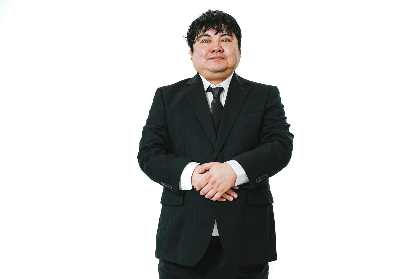 両手を体の前で組み、丁寧な姿勢で立つ黒いスーツ姿の男性［モデル：段田隼人］