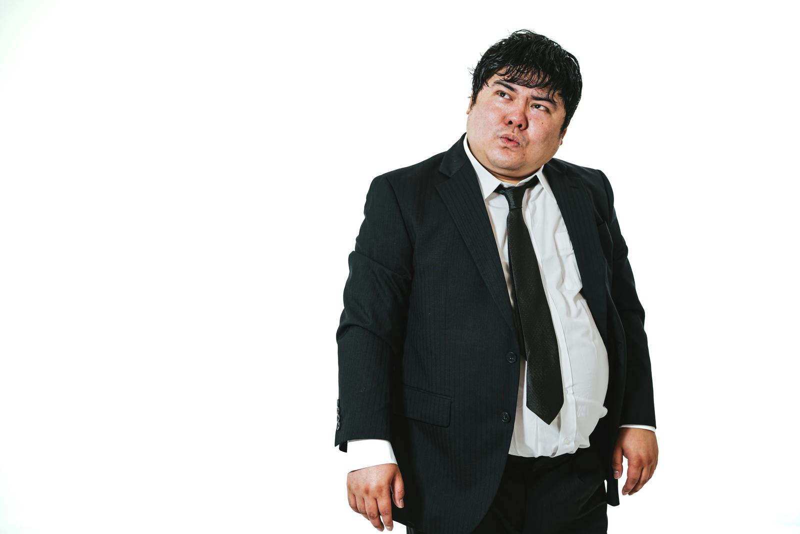 白背景で口をすぼめて上目遣いに様子をうかがう恰幅のいいスーツ姿の男性［モデル：段田隼人］