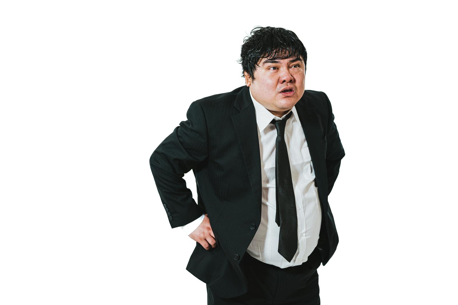 白背景の前で腰に手を当てて前かがみになり息切れしているスーツ姿の太った男性会社員［モデル：段田隼人］