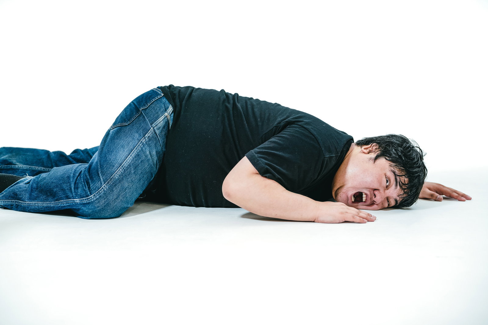 白い床に倒れ込み手で顔を覆う、デニムパンツと黒いトップスの男性［モデル：段田隼人］