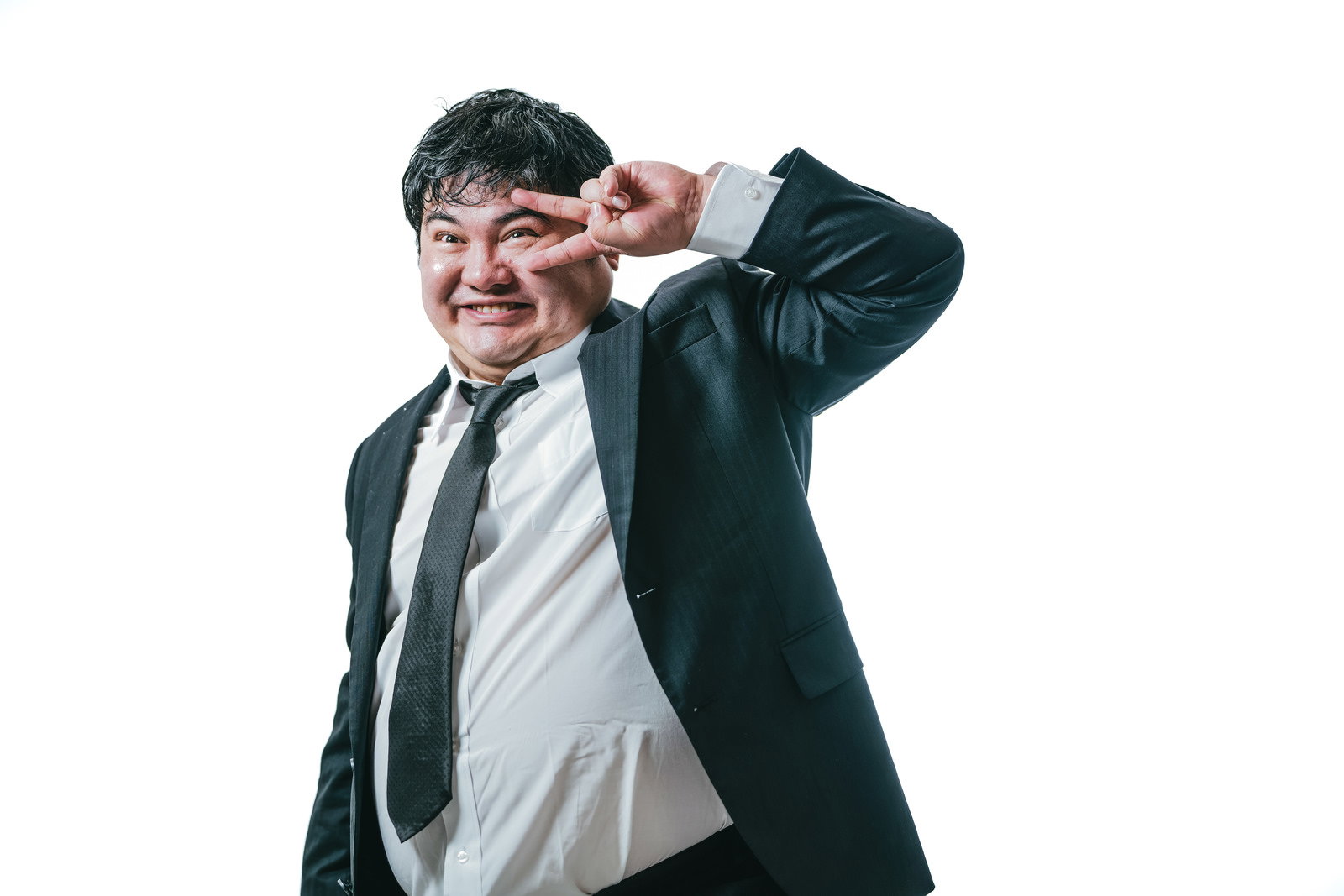 スーツ姿の男性が目の横でピースサインをして笑っている［モデル：段田隼人］