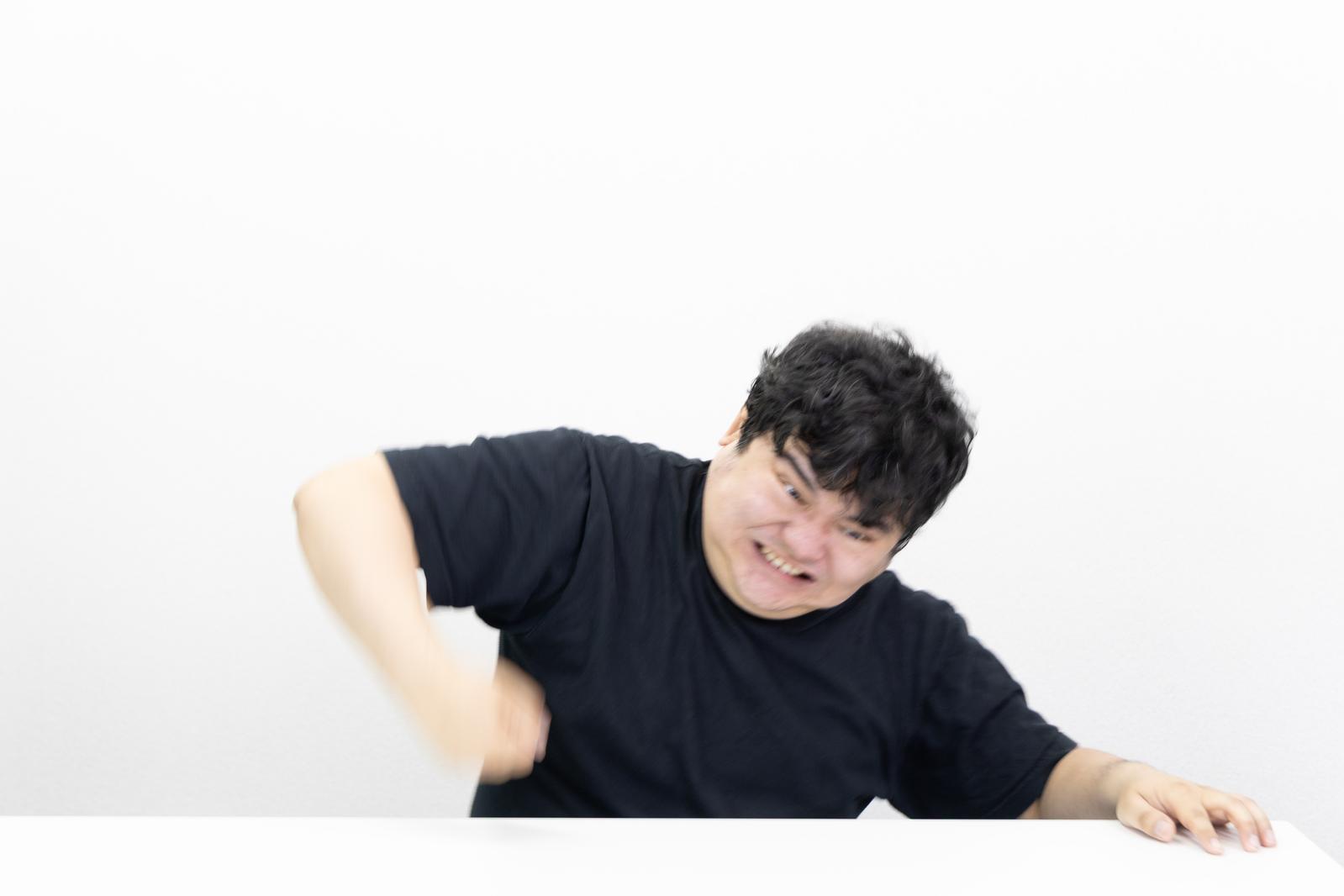 黒いTシャツの男性が怒りの表情で机を叩いている様子［モデル：段田隼人］
