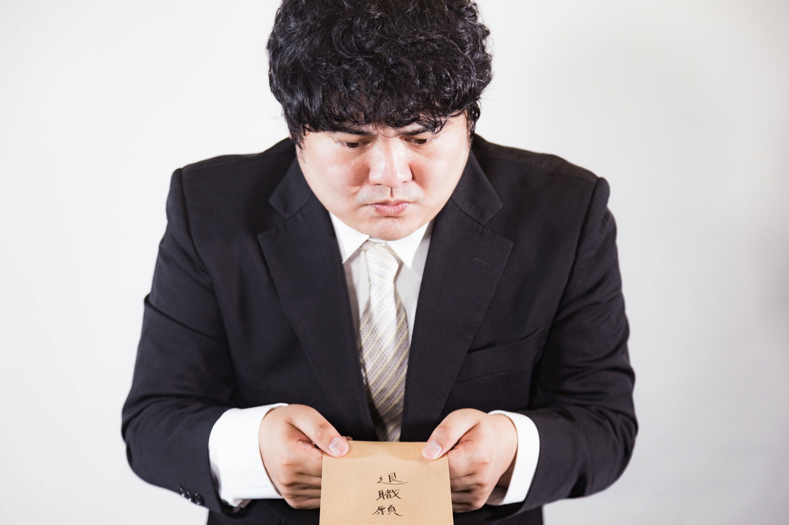 退職願を手に持ち机に向かって座る黒いスーツの男性［モデル：段田隼人］