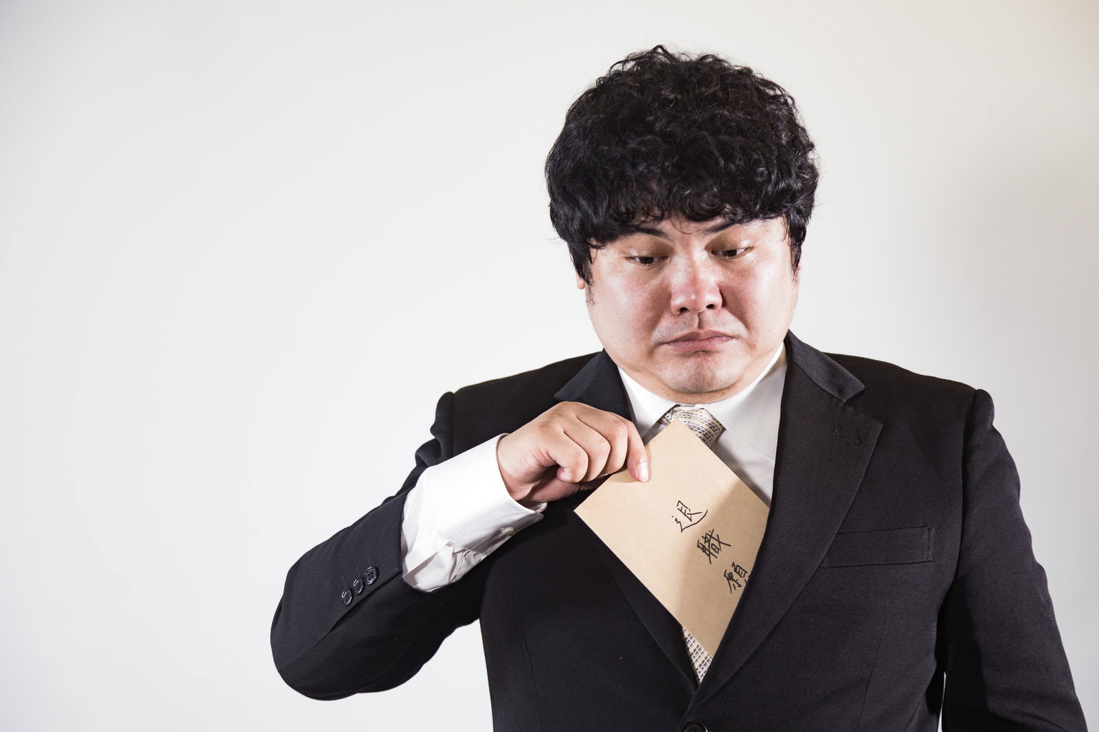 退職願を胸に抱えて不安そうな表情を浮かべる黒いスーツ姿の会社員男性［モデル：段田隼人］