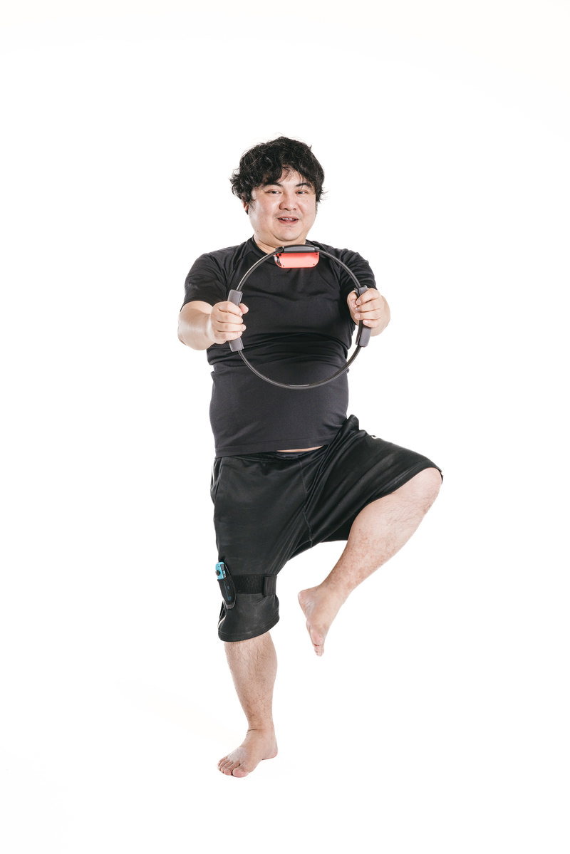 リングフィットを持ちながら片足立ちで立木ポーズを取る男性［モデル：段田隼人］
