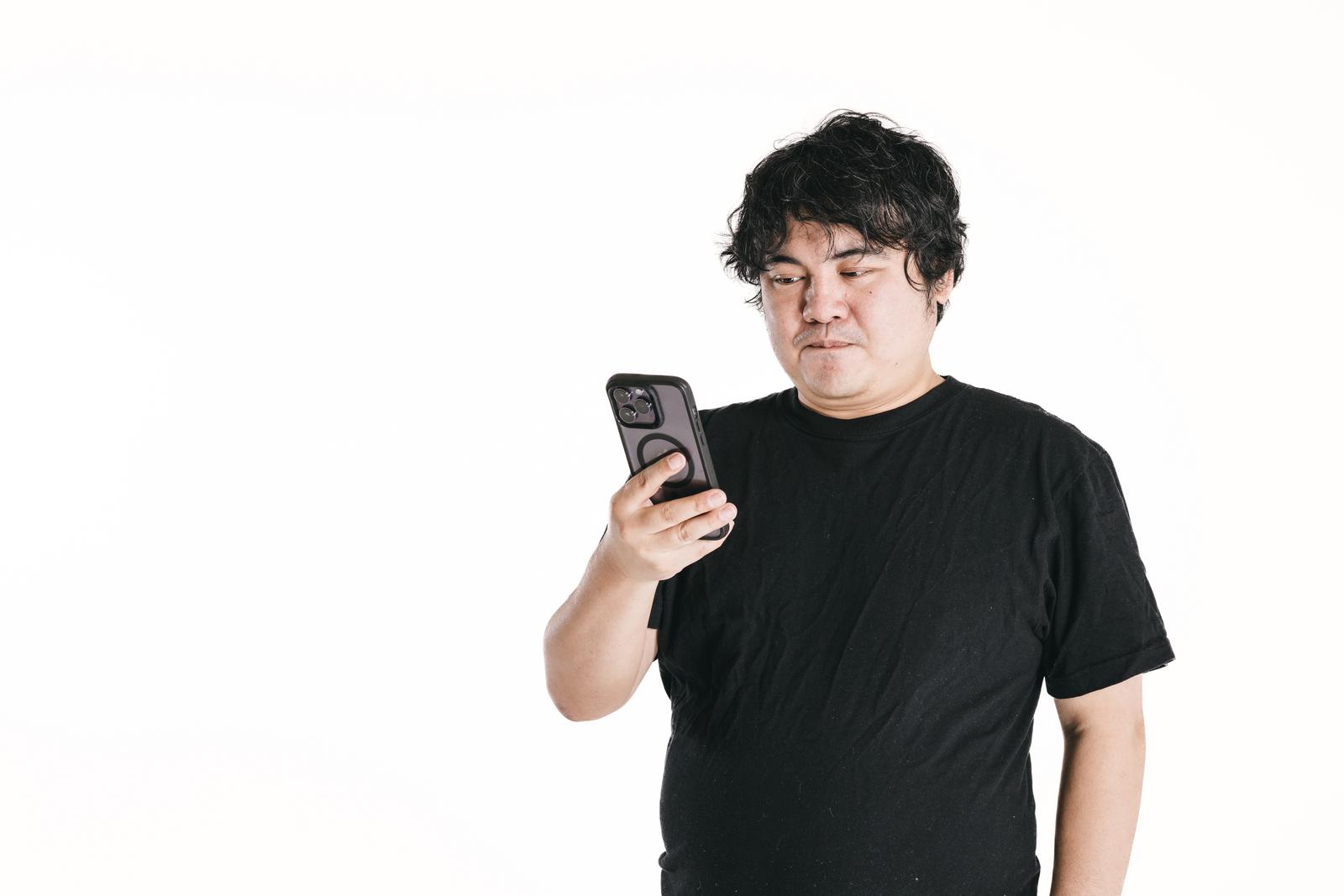 スマートフォンを持ちながら画面を見つめる不満そうな男性［モデル：段田隼人］