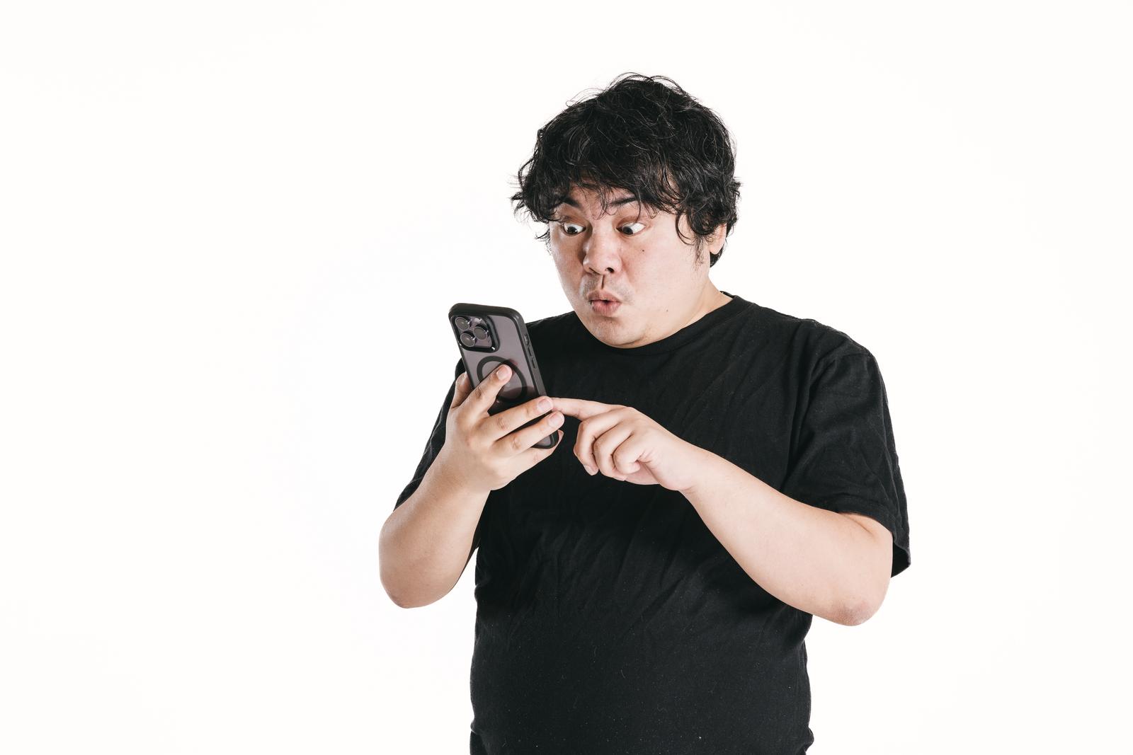黒いTシャツを着た男性がスマートフォンを両手で持ち、画面を見て目を見開いて驚いている［モデル：段田隼人］
