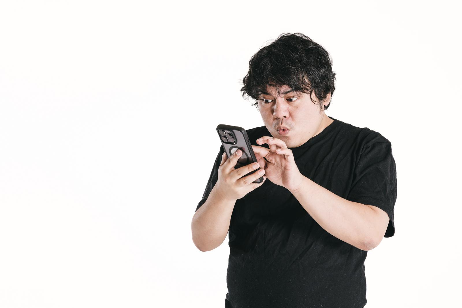 スマートフォンを凝視して驚く黒いTシャツを着た男性［モデル：段田隼人］