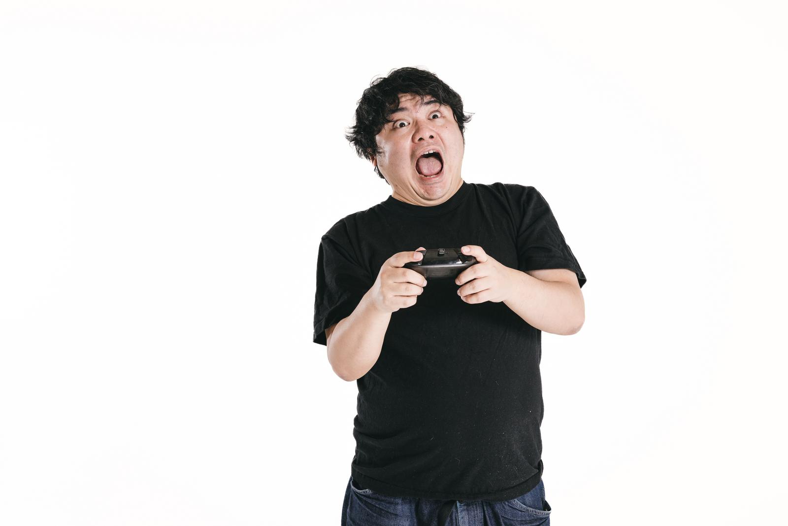 ゲームコントローラーを握りしめて驚きの表情を見せる黒いTシャツの男性［モデル：段田隼人］