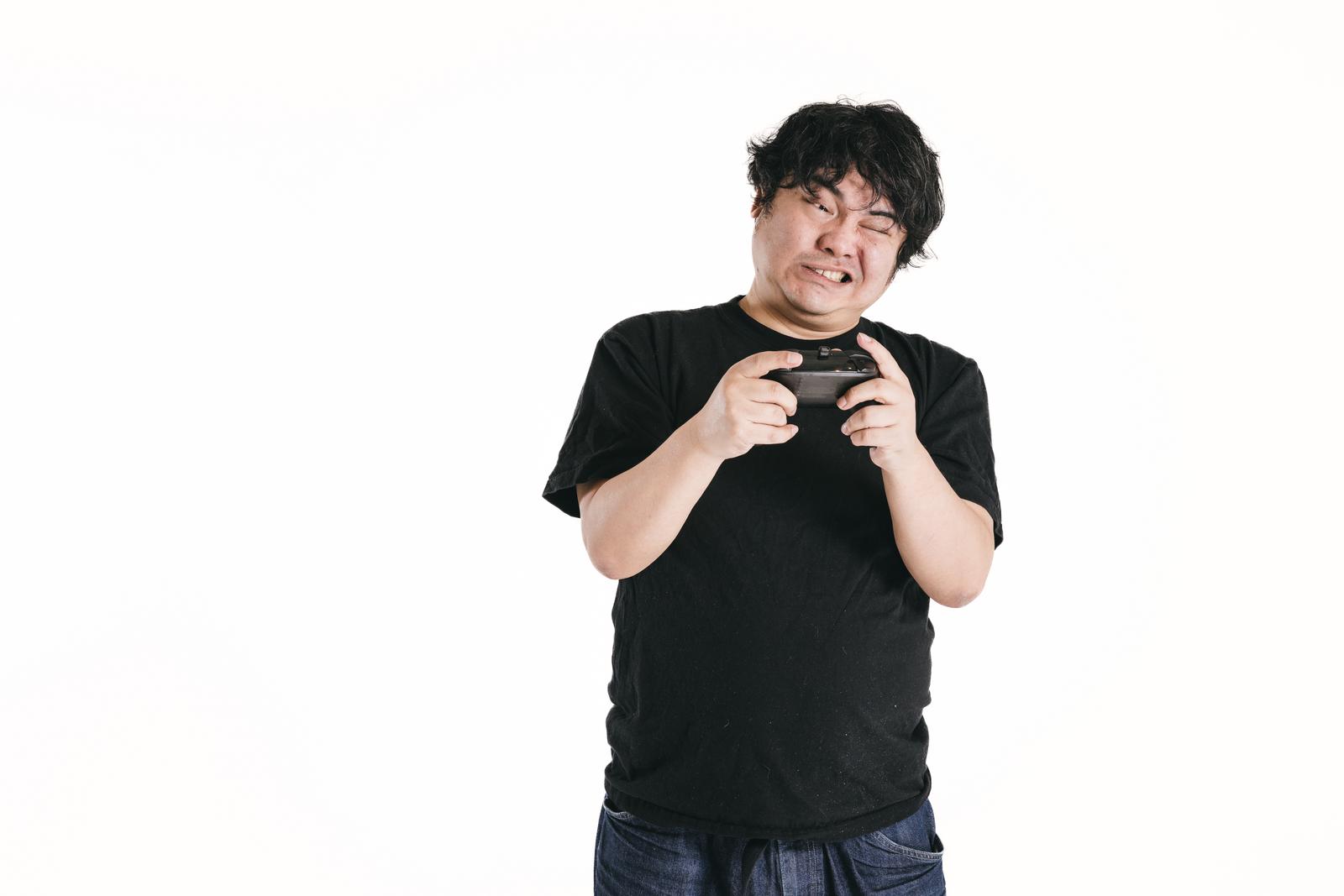 ゲームコントローラーを両手で握りしめて歯を食いしばる黒いTシャツの男性［モデル：段田隼人］