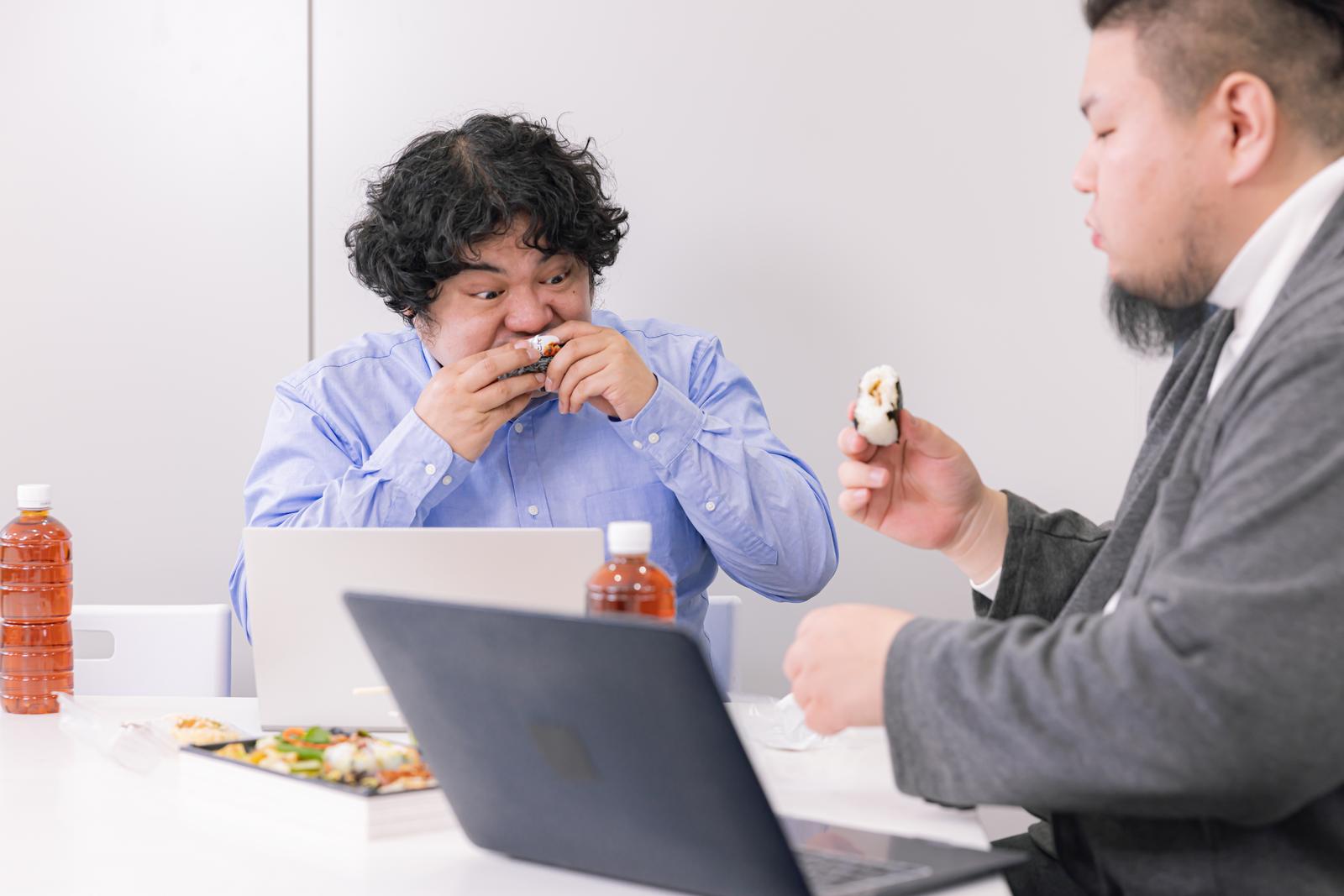 テーブルでおにぎりを食べながら打ち合わせをする2人のビジネスマン［モデル：段田隼人 うまみん］