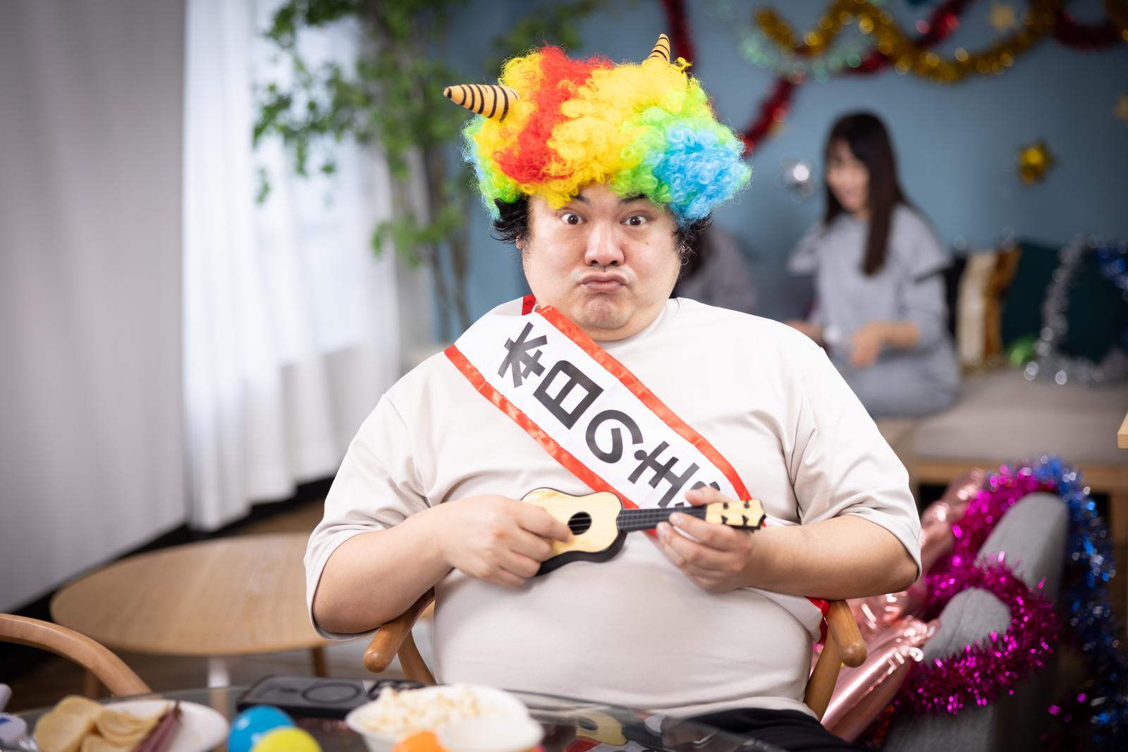 カラフルウィッグと角、たすきをつけてウクレレを持つ誕生日パーティーの男性［モデル：段田隼人］