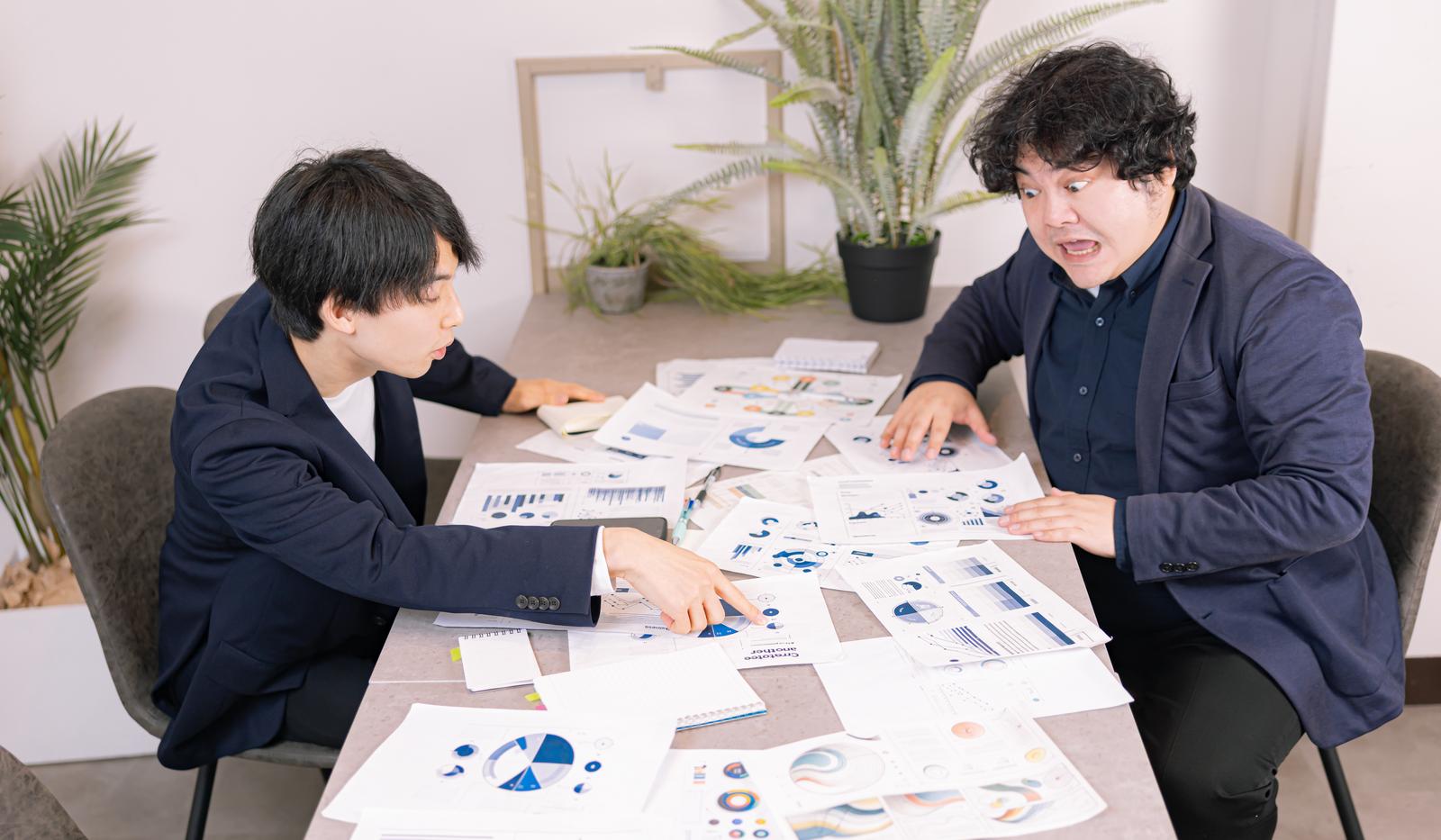 グレーのテーブルの上に資料を広げて会議するビジネスマン2人［モデル：たくみ 段田隼人］
