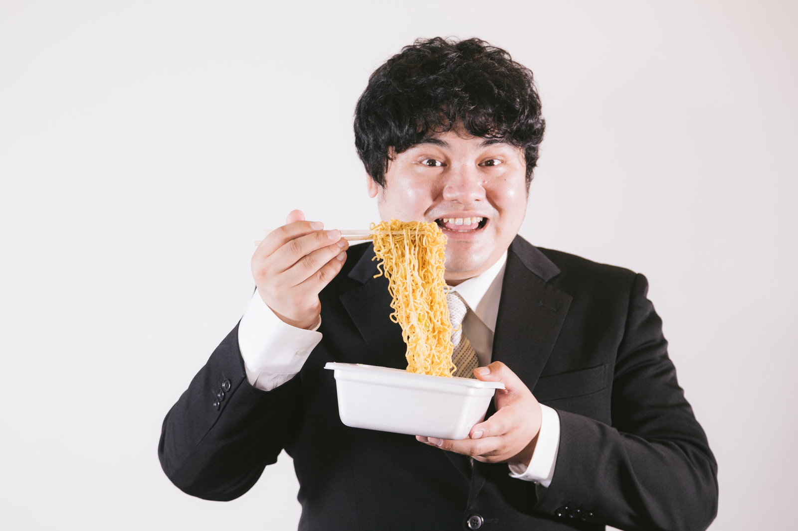 黒いスーツを着た男性が笑顔でカップ焼きそばを食べている様子［モデル：段田隼人］
