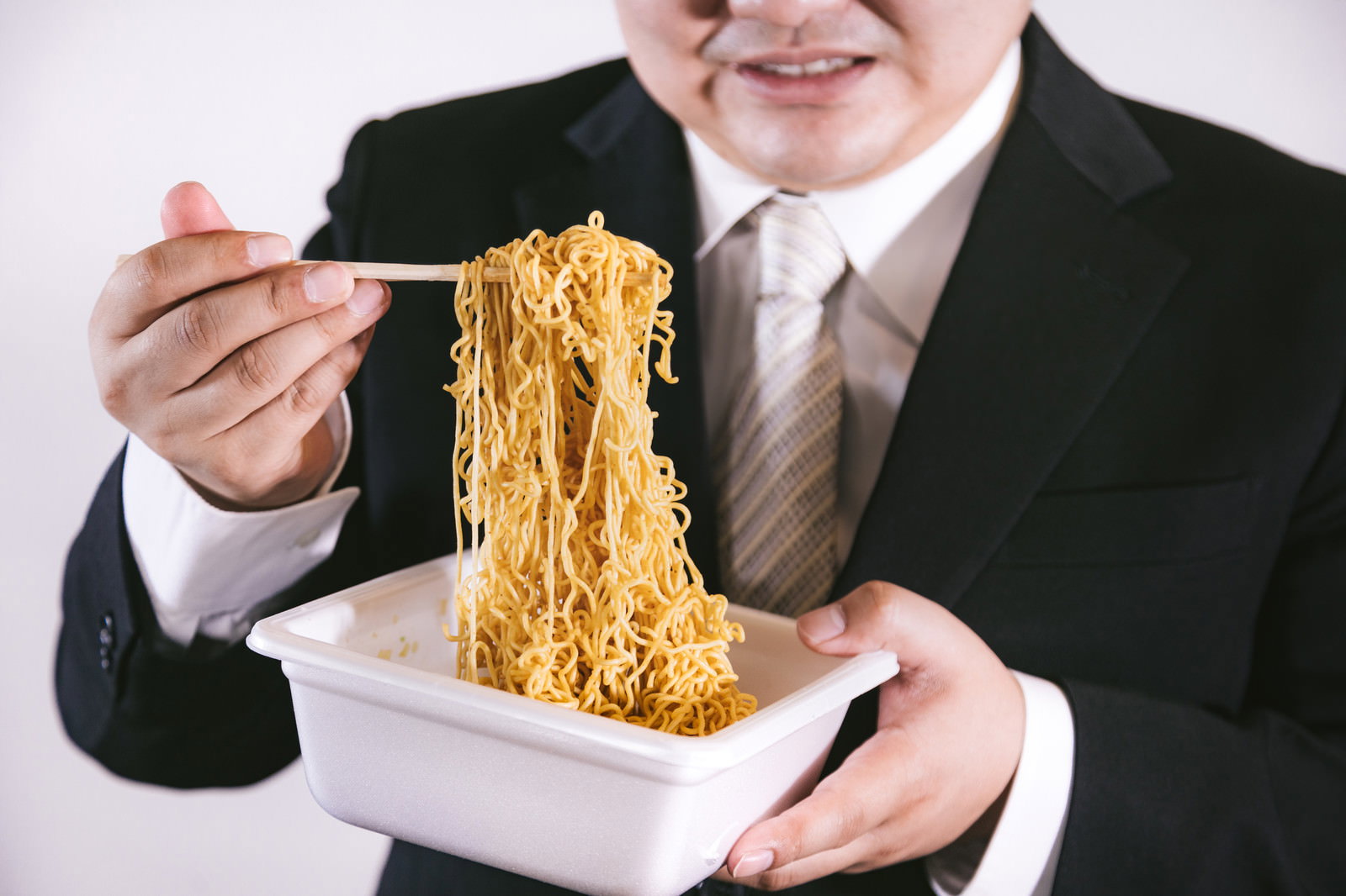 スーツを着た男性がフォークでカップ焼きそばの麺を巻き上げている［モデル：段田隼人］